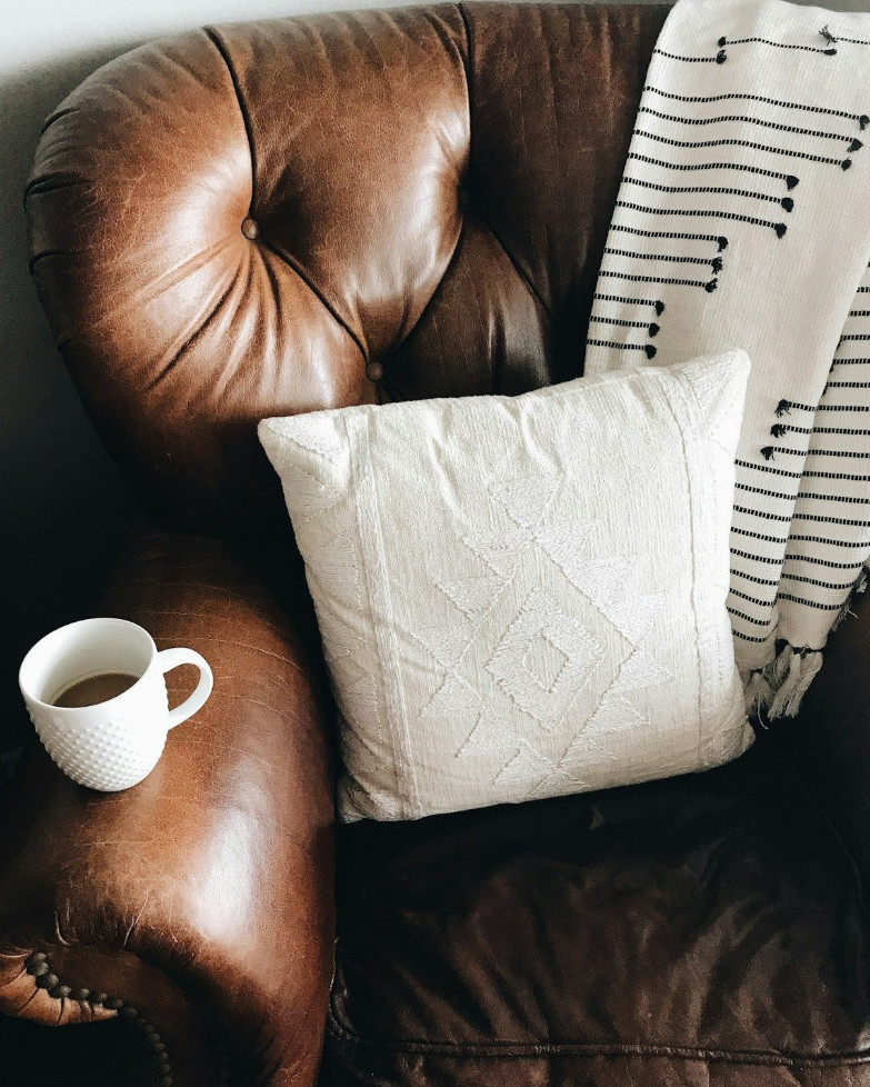 A tufted brown leather armchair with a white pillow, a striped throw and a cup of coffee on the arm